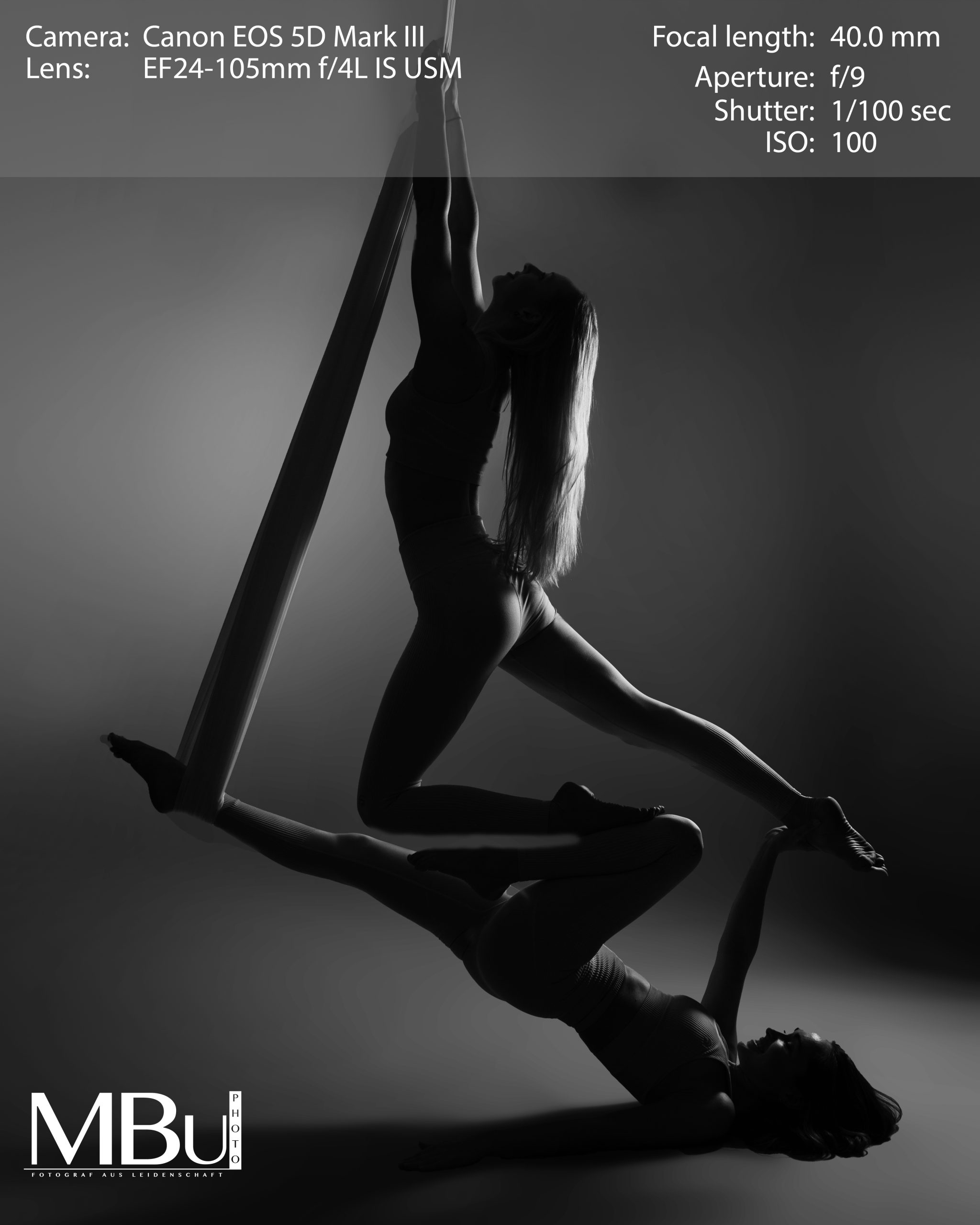Eine Aerial-Yoga Pose in Schwarz weiß. Zwei Frauen hängen in einer Yoga-Pose von einem Seil an der Decke. Eine Frau liegt mit dem Rücken auf dem Boden