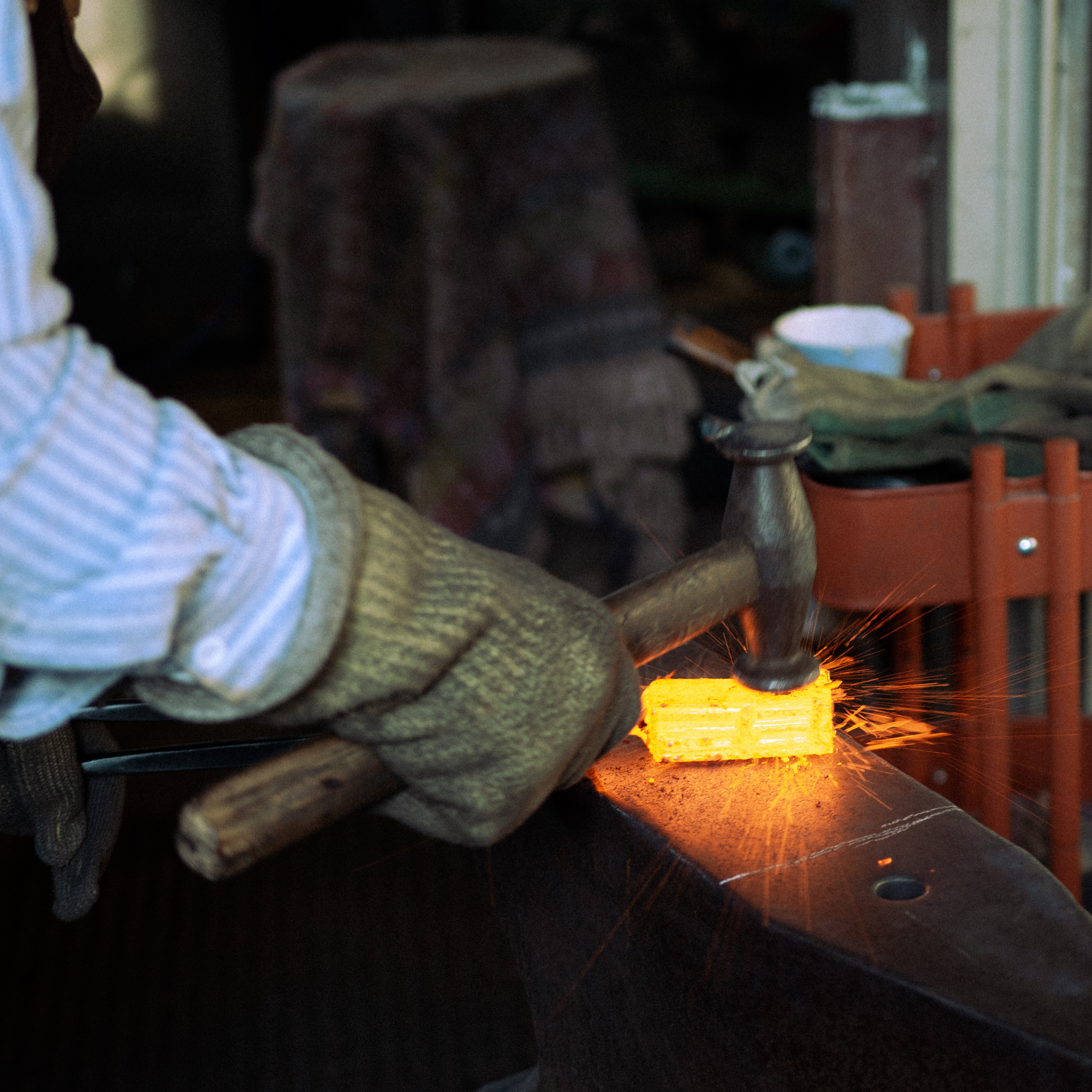 Ein Stück Stahl wird vom Schmied beschlagen. Der Stahl leuchtet heiß orange. Funken sprühen als der Hammer auf den Stahl trifft.