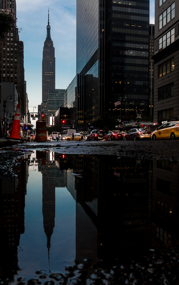 Die Straßen New Yorks im Regen. Ein Wolkenkratzer spiegelt sich in einer Pfütze.